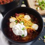 Close up of chili in a black bowl topped with cheese, sour cream, green onions, and oyster crackers.