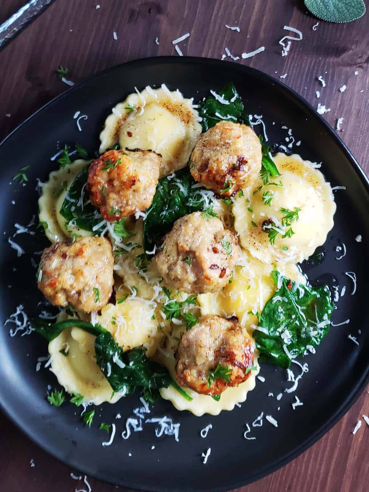 Ravioli, spinach and meatballs on a black plate.