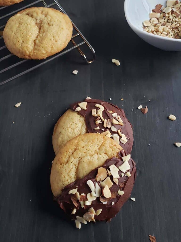 Two amaretto sugar cookies iced with melted chocolate on a dark surface surrounded by chopped almonds.