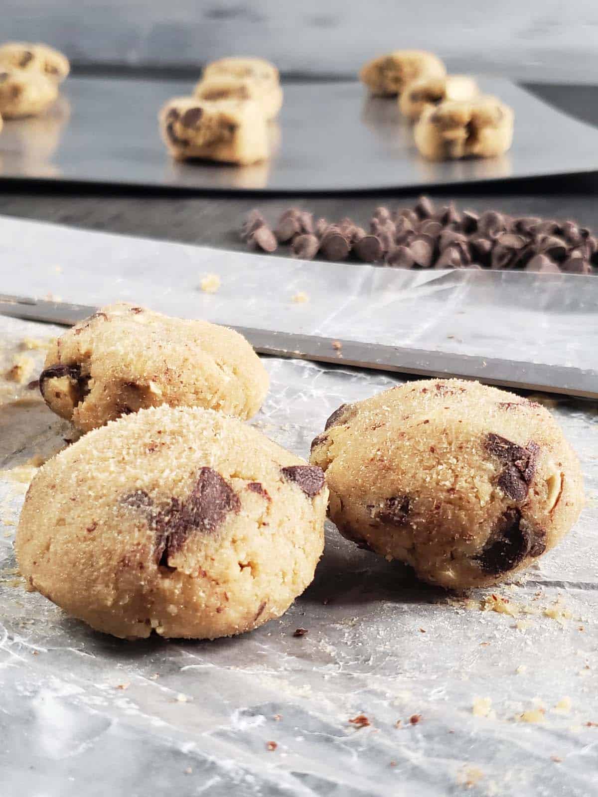 Three cookie dough balls on parchment paper with a pile of chocolate chips in the background.