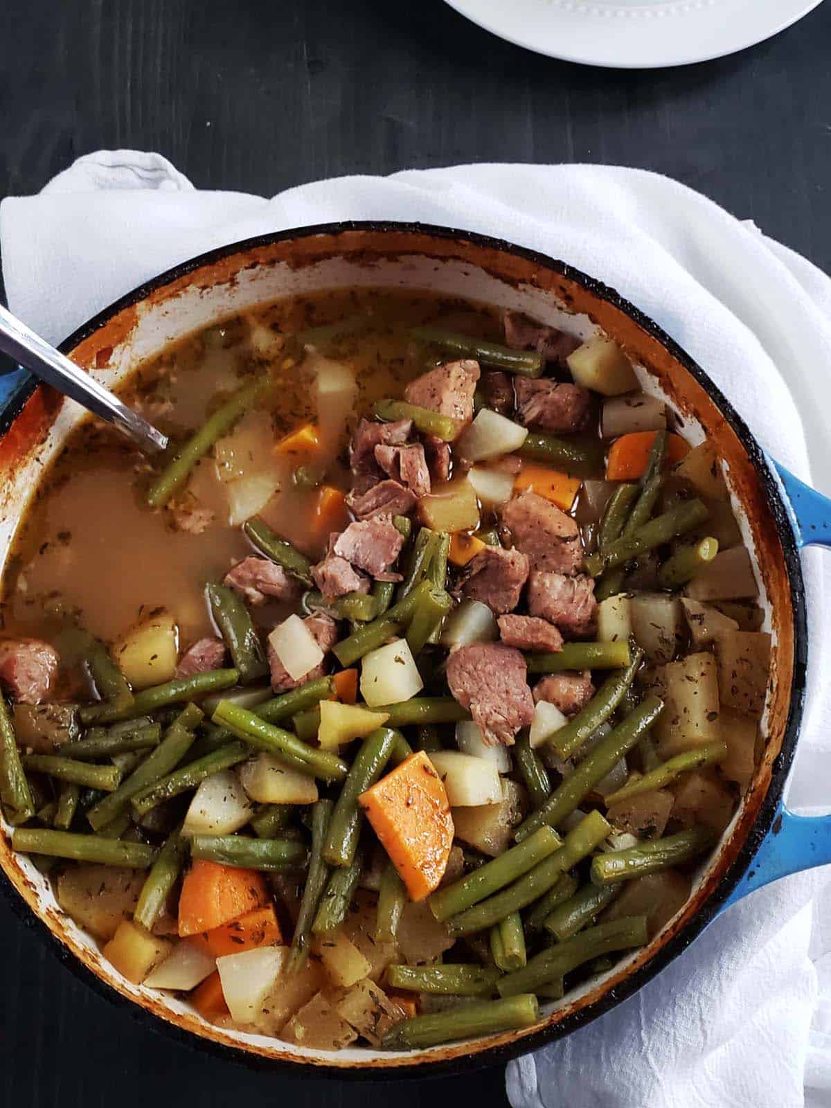 Pork stew  with green beans, sweet potatoes, apples, and kohlrabi in a blue Dutch oven.