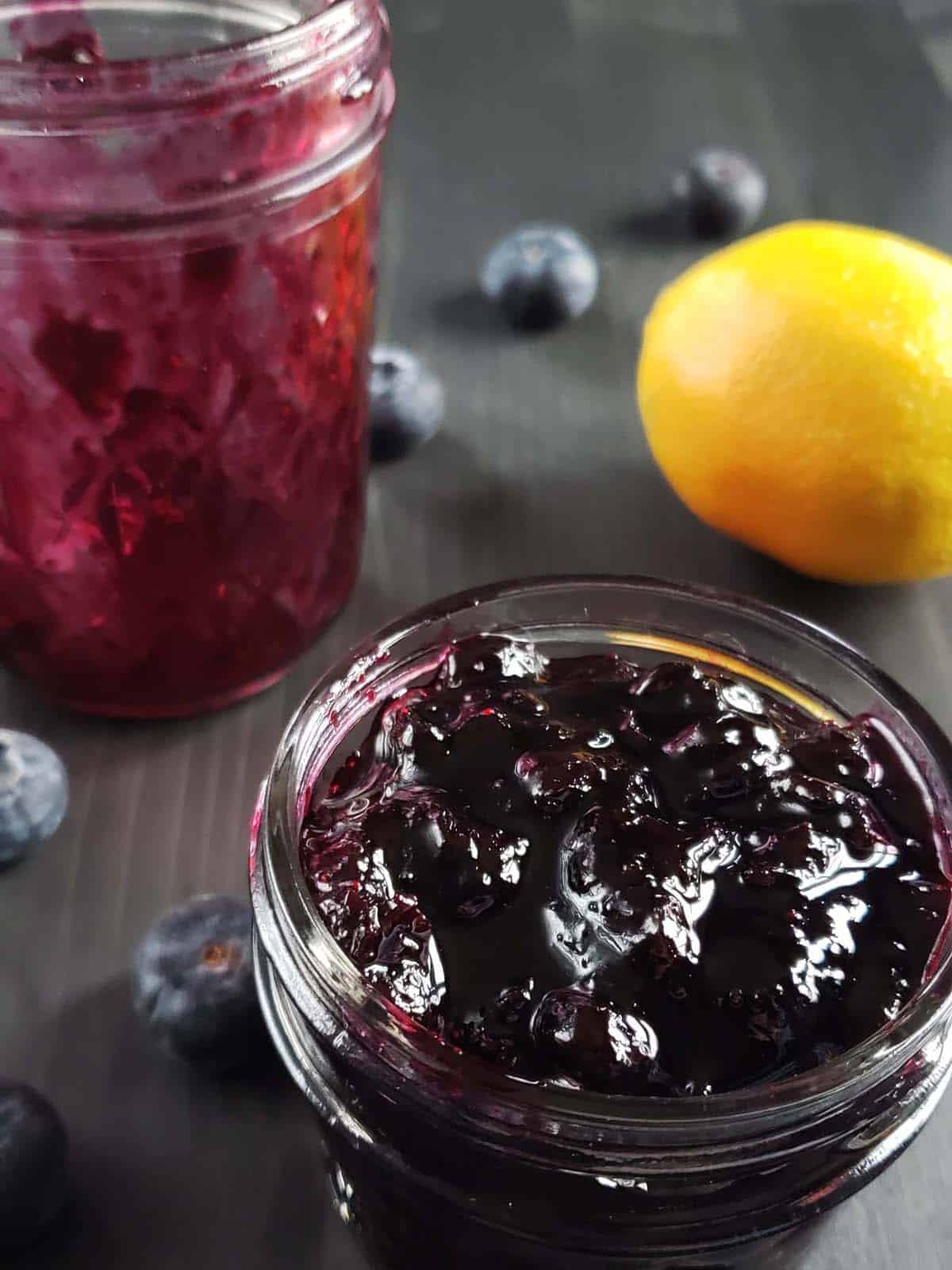 Small glass jar filled with blueberry jam surrounded by blueberries and a lemon.