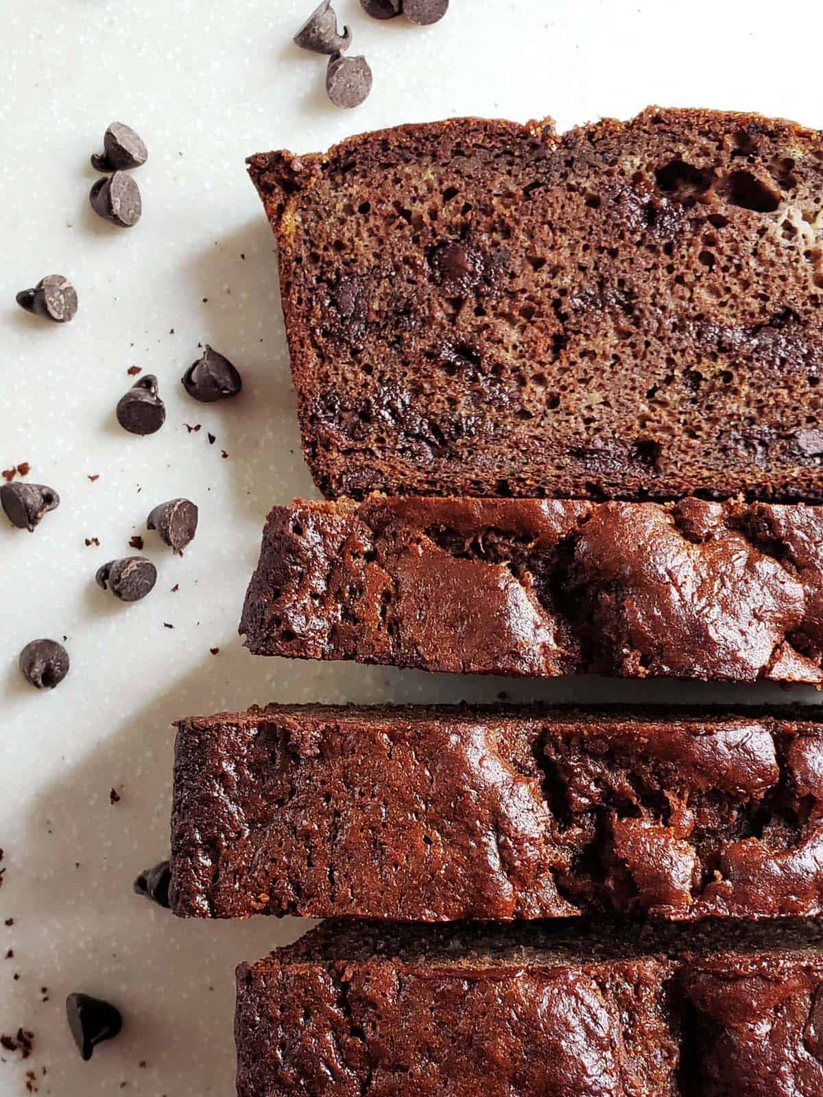 Slices of chocolate banana bread on a white surface surrounded by chocolate chips.