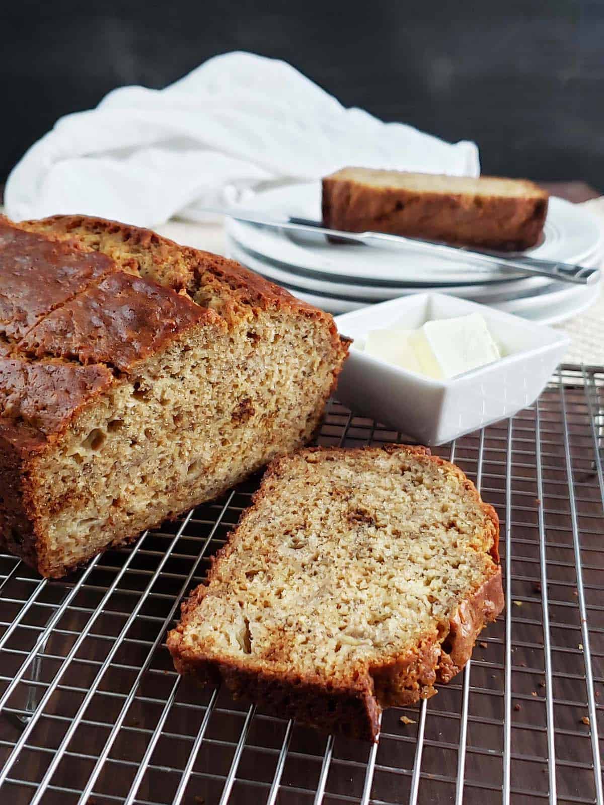 Sliced banana bread on a wire rack next to a stack of white plates and bowl filled with butter.