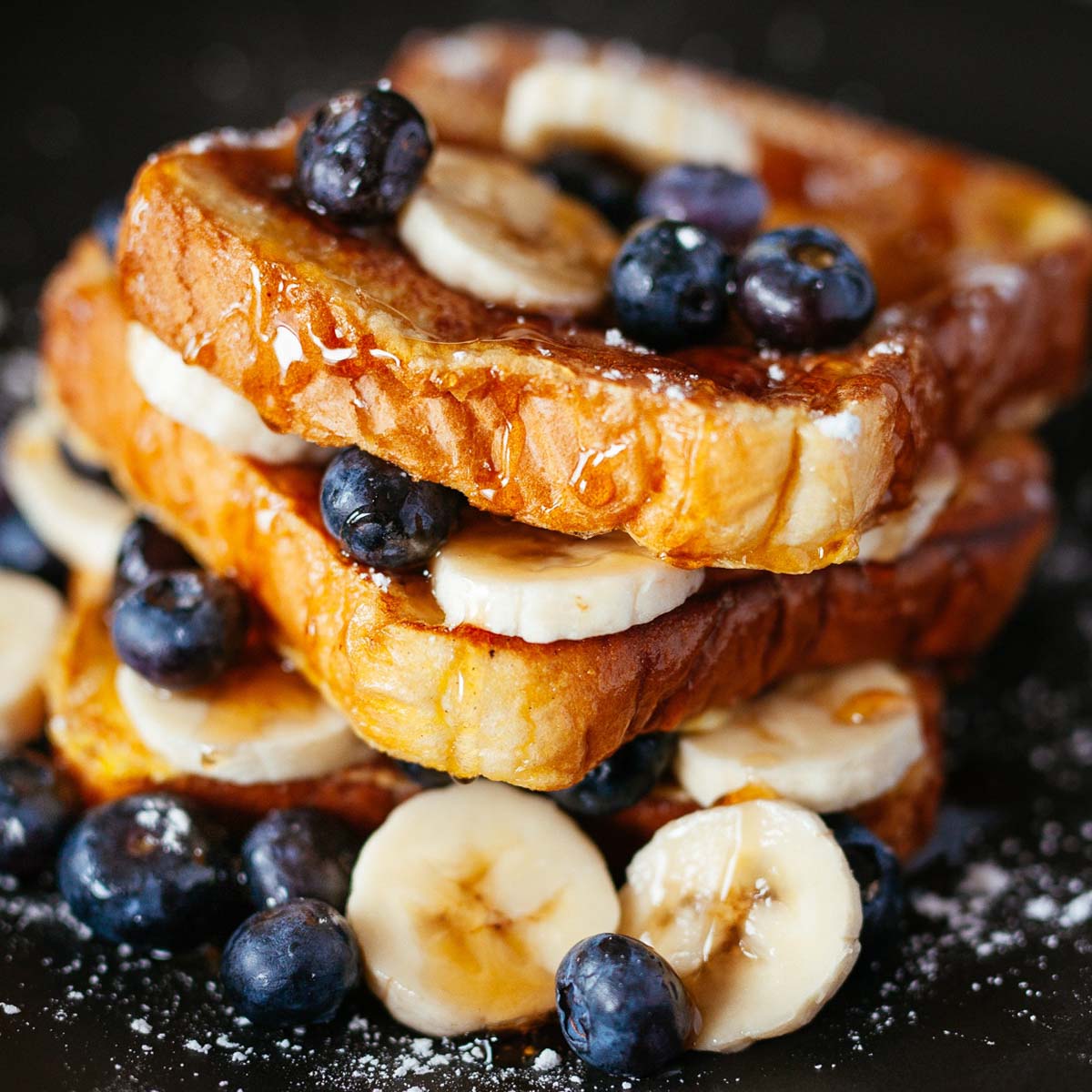 Stack of french toast on a black plate topped with blueberries and bananas.