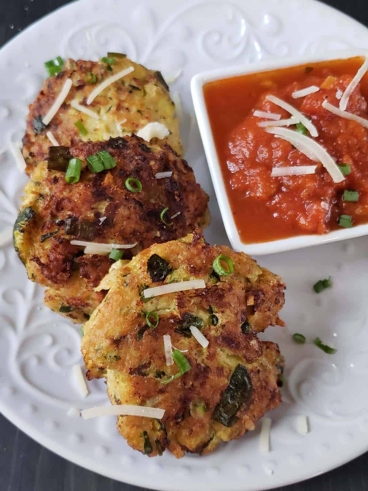 Three zucchini fritters on a white plate with marinara sauce.