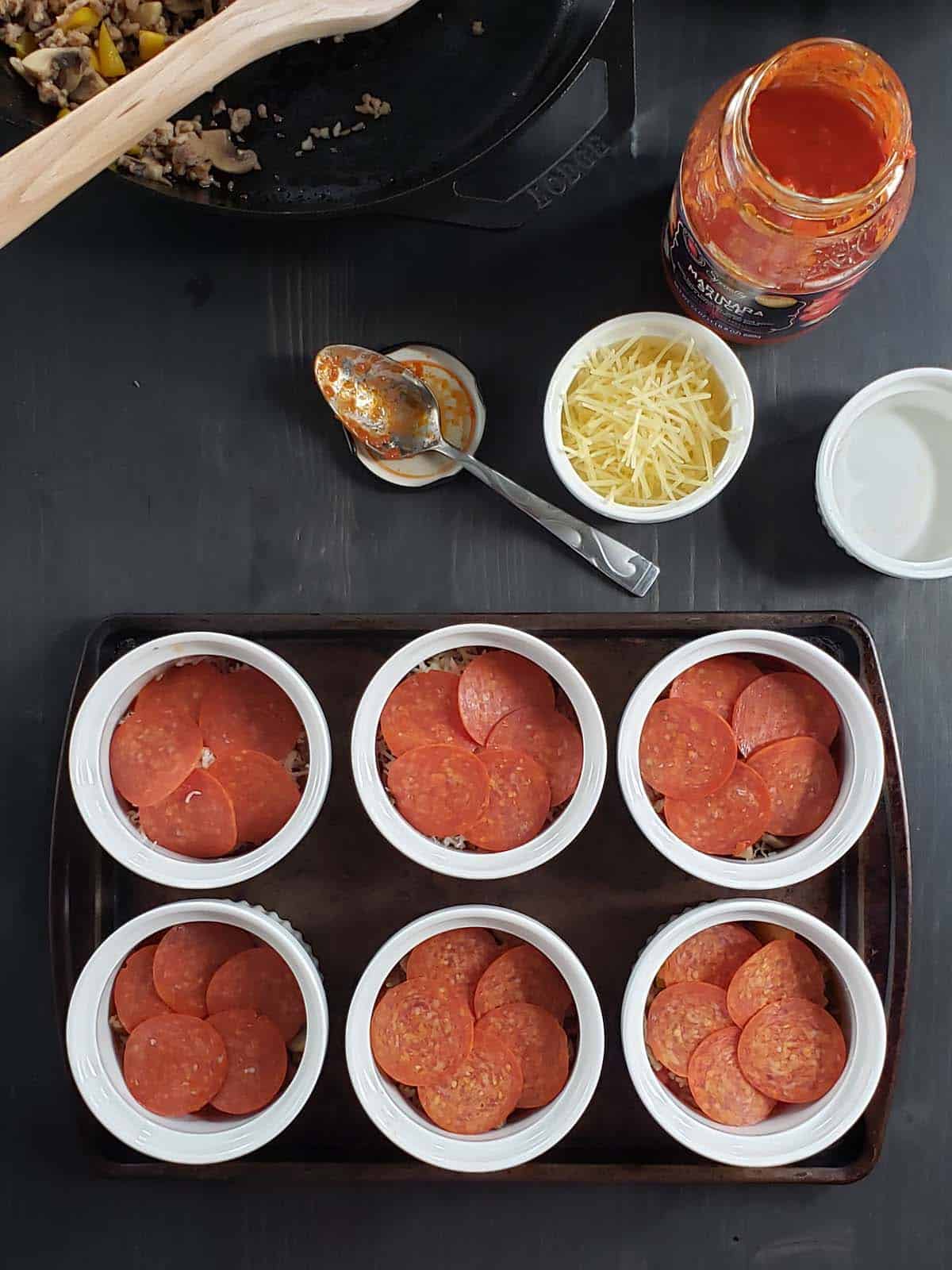 Six ramekins on a baking sheet topped with pepperoni.