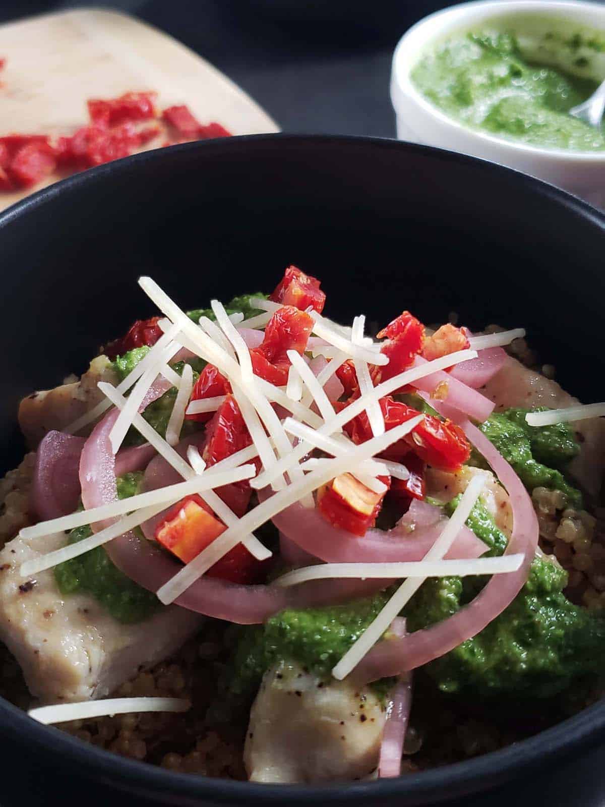 Pesto, chicken, and quinoa in a black bowl topped with pickled red onions, sun dried tomatoes, and parmesan cheese.