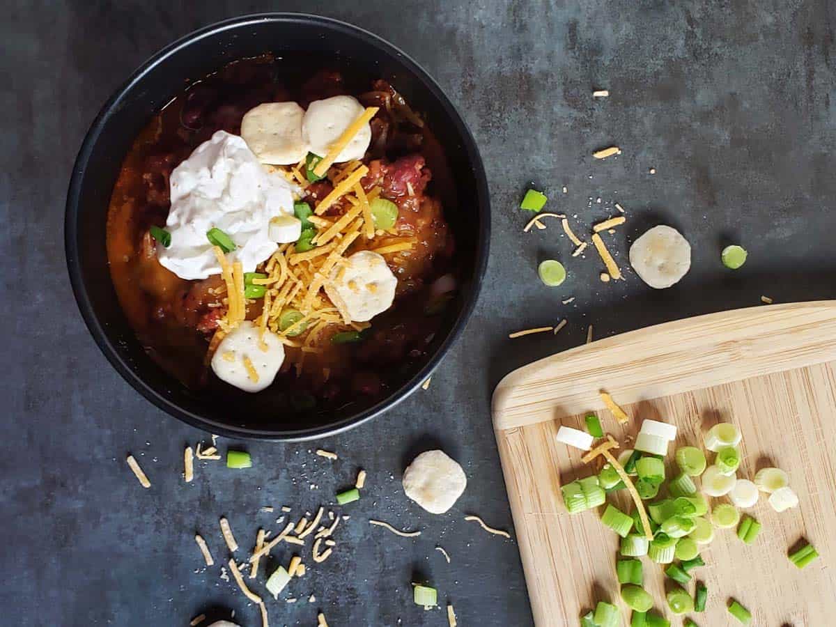 Chili in a black bowl topped with cheese, sour cream, and green onions.
