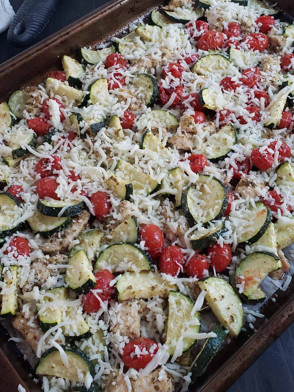 Chicken, zucchini, and tomatoes topped with cheese on a sheet pan.