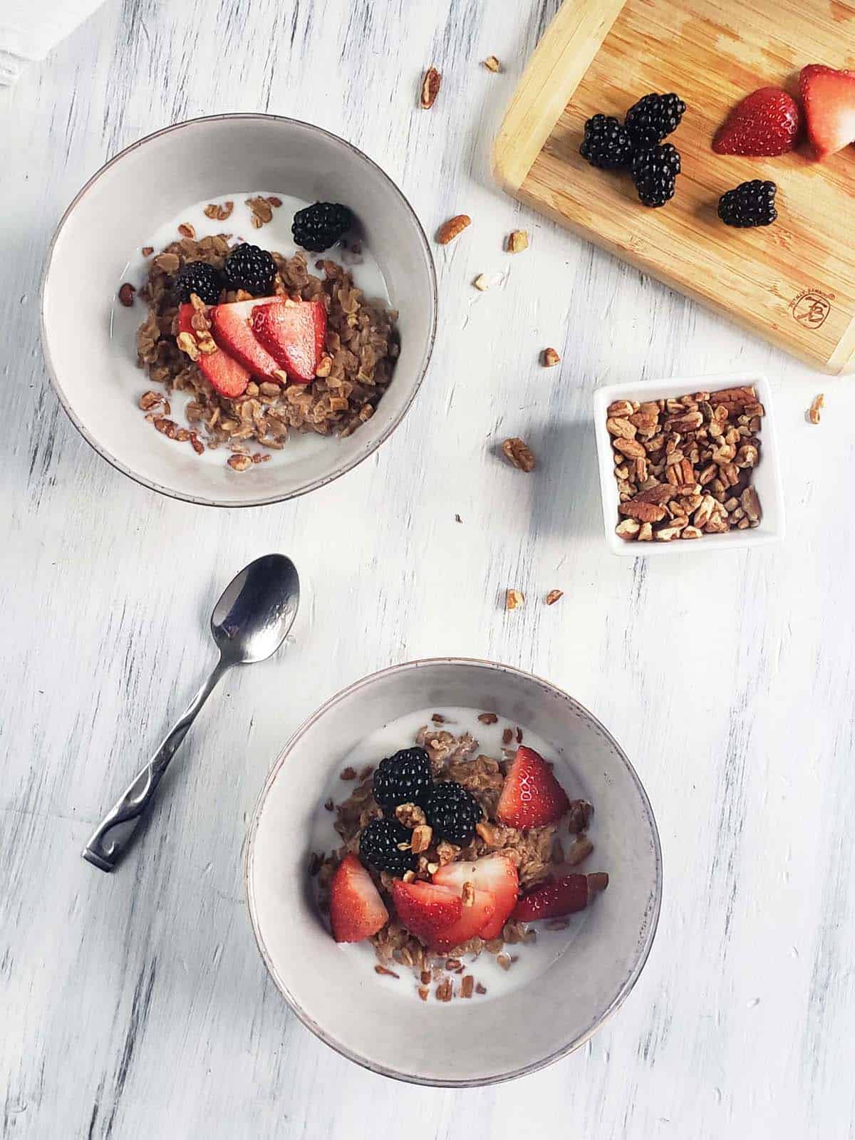 Two white bowls of oatmeal topped with berries and nuts on a white wood surface..