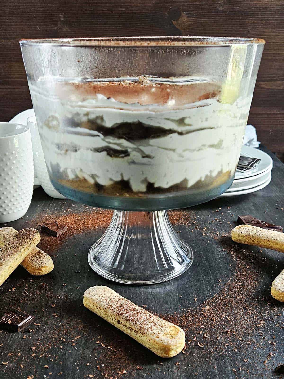 Tiramisu in a trifle dish with lady fingers and cocoa scattered around on a dark surface.