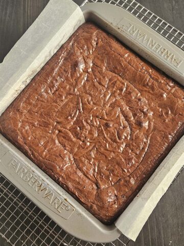 Baked brownie in a metal pan lined with parchment paper resting on a wire cooling rack.