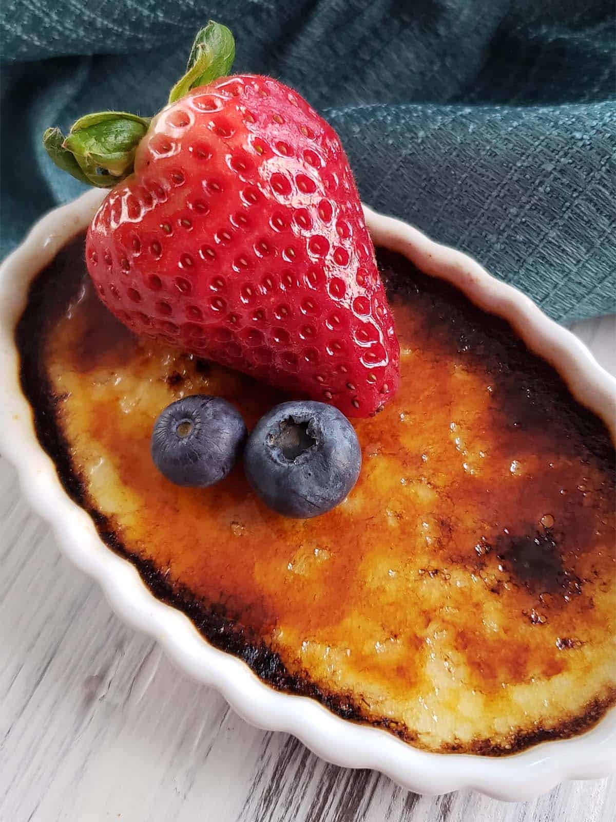Creme brulee topped with a strawberry and two blueberries on a white surface next to a blue towel.