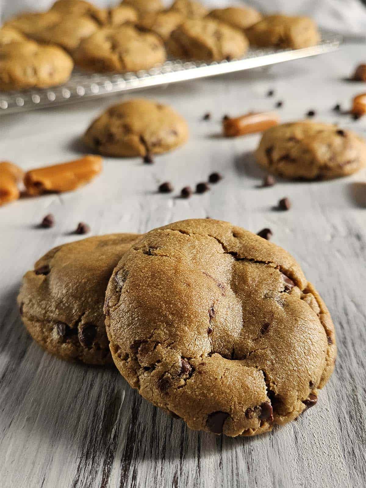 Two Peanut Butter Chocolate Chip cookies on a white wood surface surrounded by caramels and chocolate chips.