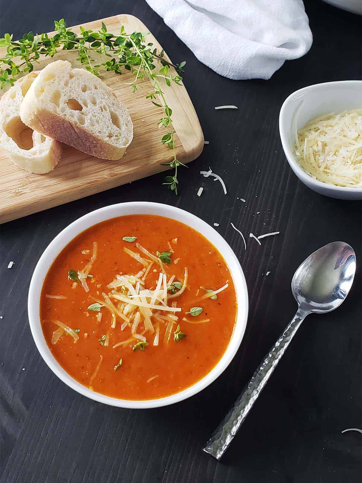 White bowl of tomato soup topped with shredded cheese and chopped herbs on a black background.